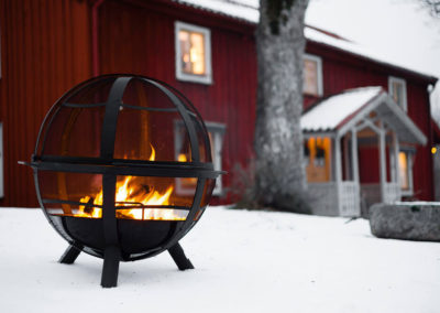 Ball shaped fire pit outside in the snow with a red house behind it